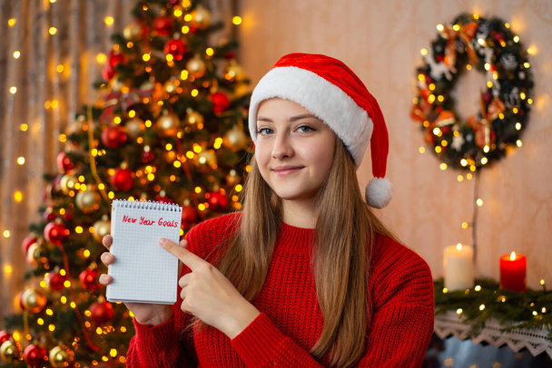 Young beautiful girl in red Santa hat with notes pointing finger to inscription New Year Goals. Concept of wish list or to do list for the comming year - Foto, afbeelding