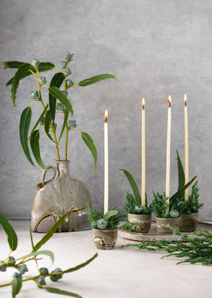 Beautiful winter floristic arrangement with white narrow candles, eucalyptus leaves and fruits in four ceramic mugs. Handmade Christmas home decoration. Selective focus. Copy space. - Valokuva, kuva