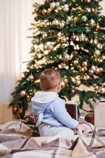 Un niño de 2 años se sienta cerca de un árbol de Navidad decorado con juguetes. Ambiente de Año Nuevo - Foto, Imagen
