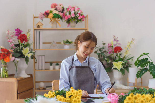 Floristry concept, Woman florist writing orders and customer information in notebook at flower shop. - Fotoğraf, Görsel