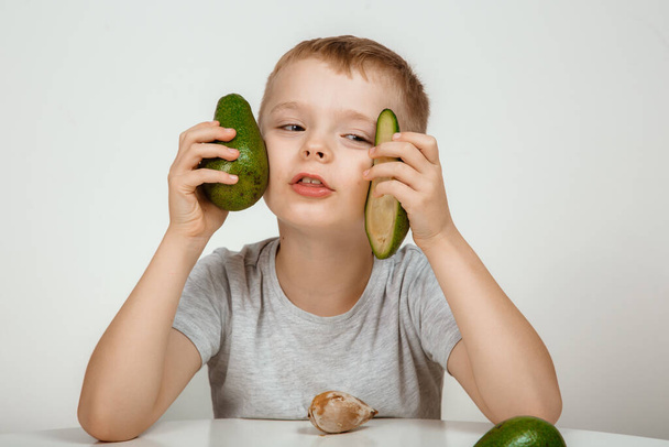 Zdravé jídlo a vegetariánství. Chlapec ukazuje avokádo a palec nahoru zblízka izolované. Avocado střih v půli. Malý kluk s čerstvým avokádem na světlém pozadí. Kvalitní fotografie. - Fotografie, Obrázek