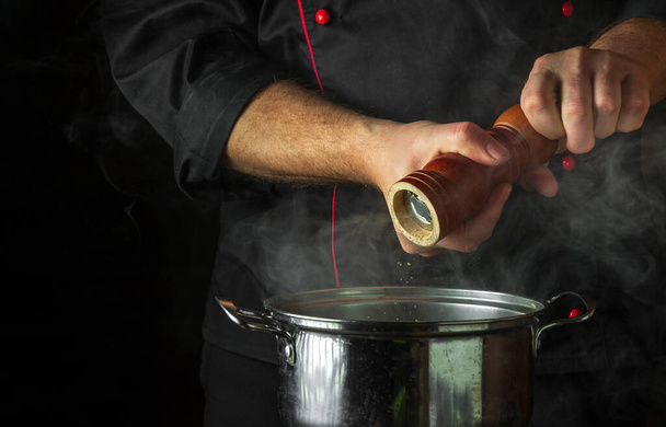 De chef voegt gemalen peper toe aan een pot kokend voedsel. Retsoran keuken koken concept met advertentieruimte op zwarte achtergrond - Foto, afbeelding