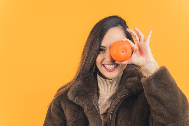 brown-haired young woman in a fur coat holding a mandarin on an eye, winter concept medium closeup isolated on orange background. High quality photo - Photo, Image
