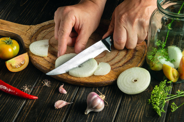 De chef snijdt de ui met een mes op een keukensnijplank voor conservering in een pot. Heerlijke conserven groenten door de handen van de kok - Foto, afbeelding