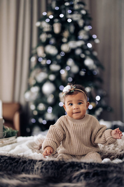 幸せな混合レース赤ちゃんの女の子はクリスマスツリーの背景にベッドの上に座っている。異人種間の家族と団結の概念. - 写真・画像