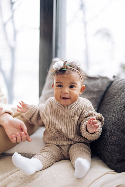 Happy mixed race baby girl sits near pillows against window. Baby has wreath with Christmas decoration on head. Concept of interracial family and unity between different human races. - Photo, Image
