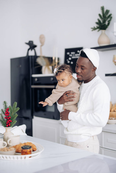 幸せなアフリカの男は皿の上のクリスマスの装飾そしてペストリーの近くの台所で彼の混合レースの赤ん坊の娘を保持する。異人種間の家族と団結の概念. - 写真・画像