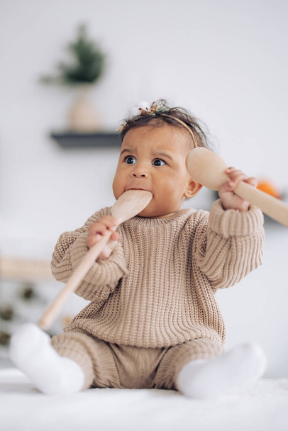 Cute mixed race baby girl sits on table in kitchen and plays by wooden spoons. Baby has wreath with Christmas decoration on head. Concept of interracial family and unity between different human races. - Fotografie, Obrázek