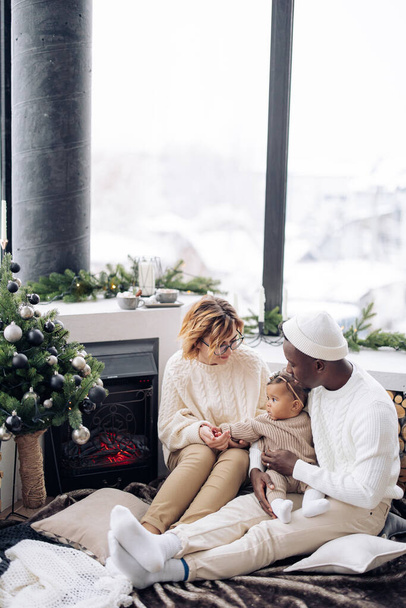 赤ちゃんの娘と幸せな異人種間の家族は、クリスマスツリーと装飾を背景に暖炉の近くに座っています。異人種間の家族と団結の概念. - 写真・画像