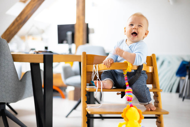 Onnellinen pikkulapsi istuu ja leikkii lelullaan perinteisessä skandaalimaisessa puisessa syöttötuolissa modernissa kirkkaassa kodissa. Söpö vauva. - Valokuva, kuva