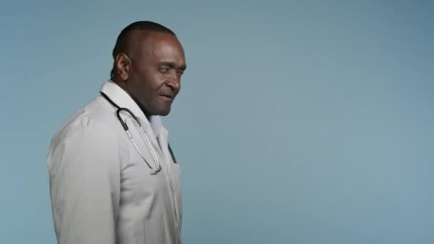 Afrikkalainen amerikkalainen mies lääketieteen puku kävely harmaa tausta studio mies lääkäri sairaalan työntekijä kutsuu potilaita käyttämään ammatillista palvelua medicare heiluttaa käsi tekee tänne kutsuva ele - Materiaali, video