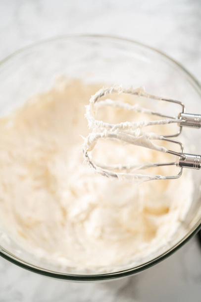 Whisking buttercream frosting in a glass mixing bowl with an electric hand mixer to prepare the peppermint buttercream frosting. - 写真・画像