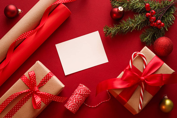 Χριστουγεννιάτικα στολίδια, δώρα και διακόσμηση με λευκό κενό χαιρετισμό ή επαγγελματική κάρτα ή να σας ευχαριστήσω σημείωμα στο εορταστικό κόκκινο φόντο ως χαρούμενες διακοπές flatlay. - Φωτογραφία, εικόνα