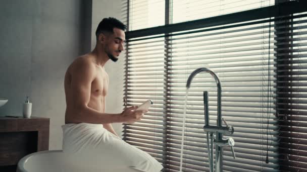 Bain du matin dans la salle de bain hygiène indien hispanique nu homme sexy gars assis sur la baignoire avec serviette sur les hanches avant de se laver tenant gel douche baume shampooing pour lotion capillaire huile corporelle crème cosmétique masculin - Séquence, vidéo