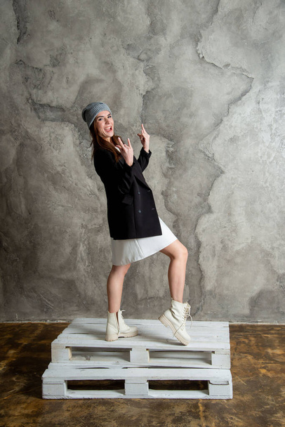 beautiful young woman posing in studio wearing white dress, black jacket and gray hat - Фото, изображение