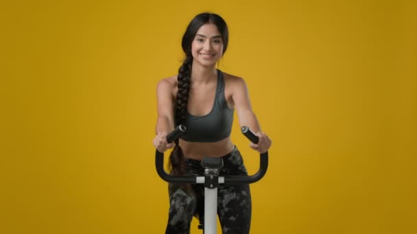 Индийская стройная счастливая улыбчивая спортивная женщина спортивная девушка-велосипедистка велосипедистка гибкая езда на велосипеде спортивное снаряжение фитнес-тренировки кардио ездить в желтой студии большой палец вверх жестом - Кадры, видео