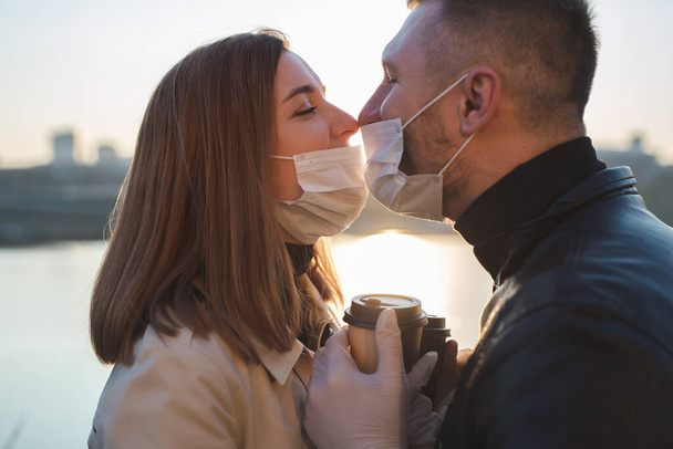 Jonge echtparen, een man en een vrouw met medische maskers en handschoenen, drinken koffie uit wegwerpbekers op straat en kijken elkaar aan tegen de achtergrond van de rivier en de stad. Quarantaine. - Foto, afbeelding
