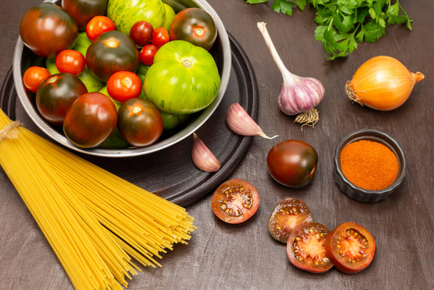 Green and red tomatoes in bowl. Pasta, chopped tomatoes, onion and garlic on table. Top view. Brown background. - Φωτογραφία, εικόνα