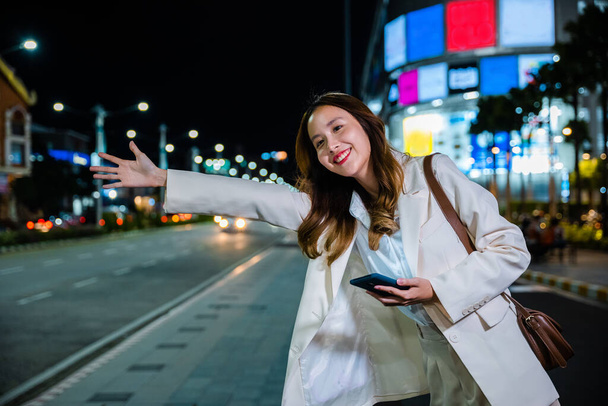 Asian businesswoman standing hail waving hand taxi on road in busy city street at night, beautiful woman smiling using mobile phone application hailing with hand up calling cab after late work - Фото, изображение