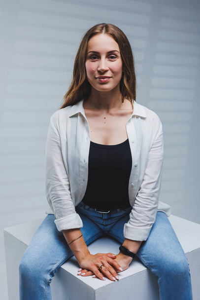 Young smiling woman in a white shirt, black t-shirt and jeans on a gray background. Portrait of a brunette woman with a smile on her face - Photo, image