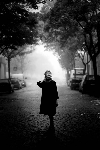 Μια κομψή γυναίκα με φθινοπωρινό παλτό στέκεται έξω τυλιγμένη στην πρωινή ομίχλη. Ασπρόμαυρη φωτογραφία. - Φωτογραφία, εικόνα