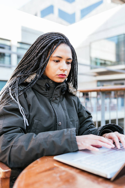 Hispanic woman with Afro braids in warm jacket sitting at table on balcony and working on laptop remotely - Zdjęcie, obraz