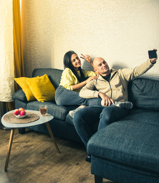 Pareja Utilice el dispositivo Smartphone, Sentado en el sofá acogedor apartamento. Tomar selfie juntos como novio y novia. Hacer chat de vídeo en Internet, Uso de las redes sociales, Streaming de vídeo en vivo - Foto, Imagen