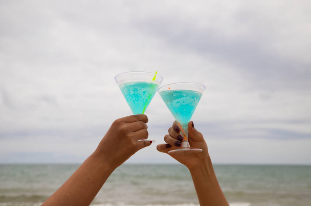 Detail of two hands raising glasses in the air. They are toasting for the good things that happen. The photo is taken from below and you can see the sky and the sea in the background. - Фото, изображение