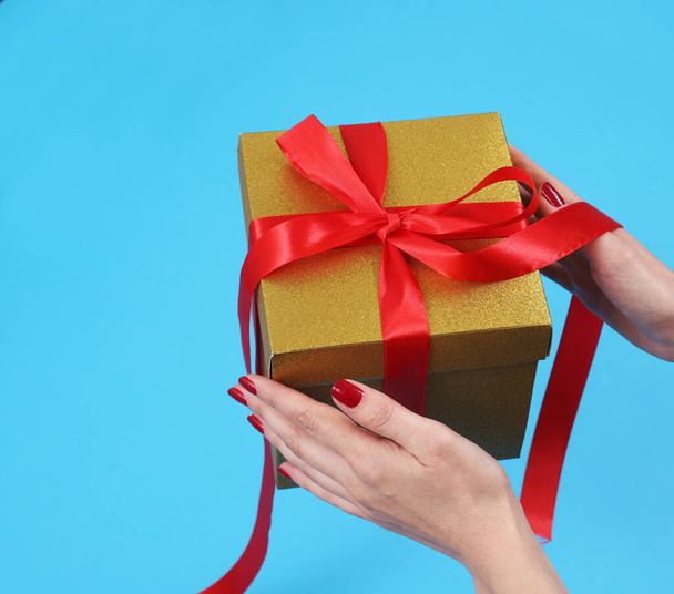 Жіночі руки розпаковують золоту крафт-паперову подарункову коробку з бантом червоної стрічки на ізольованому синьому фоні
 - Фото, зображення