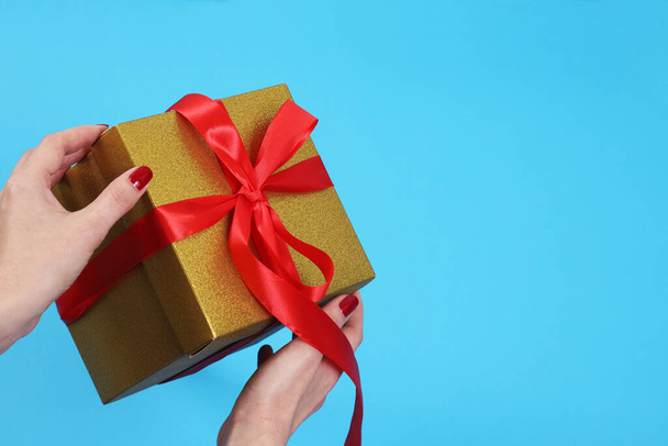Female hands unpacking golden kraft paper gift box with red ribbon bow on isolated blue background - Foto, imagen