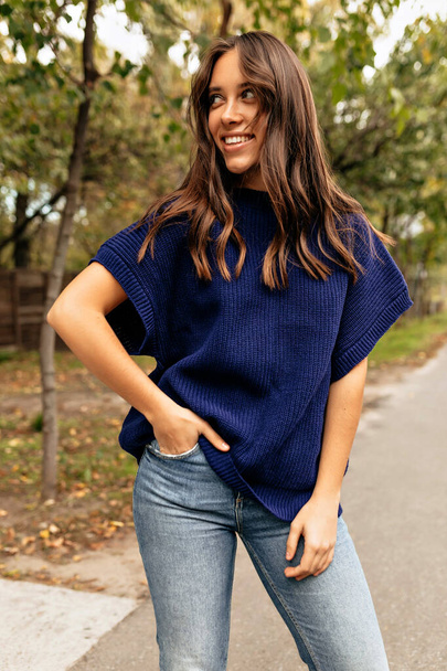 Outdoor photo of smiling attractive woman with happy smile looking aside and standing on background of green park in the city. She wears knitted shirt and jeans  - 写真・画像