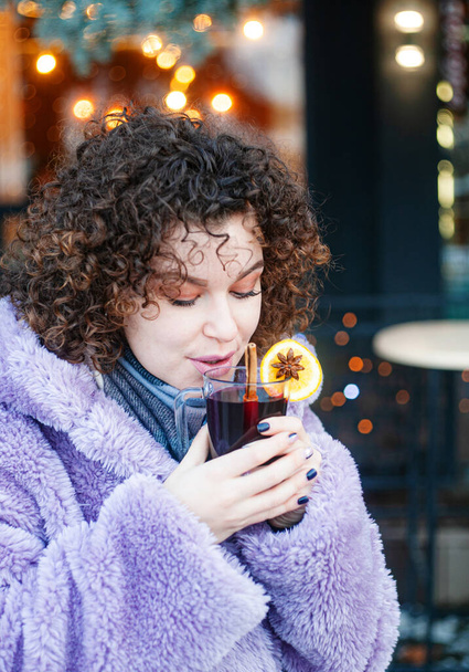 encaracolado mulher em bebidas casaco violeta na moda vinho mulled ao ar livre, fundo festivo  - Foto, Imagem