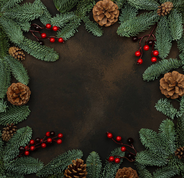 Close up border frame of fresh green spruce Christmas tree branches with cones and red holly berries decoration, over dark brown background with copy space - Фото, изображение