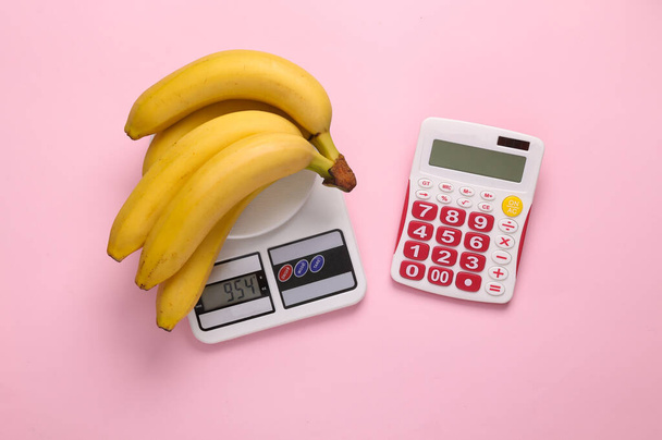 Banany na wadze i kalkulator na różowym tle. Liczenie kalorii, zdrowy tryb życia - Zdjęcie, obraz