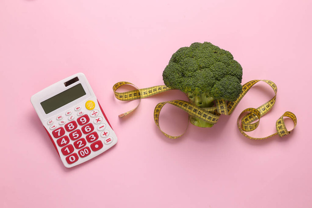 O conceito de contagem de calorias, perda de peso, dieta. Brócolis envolto com uma fita métrica e uma calculadora em um fundo rosa. Vista superior - Foto, Imagem