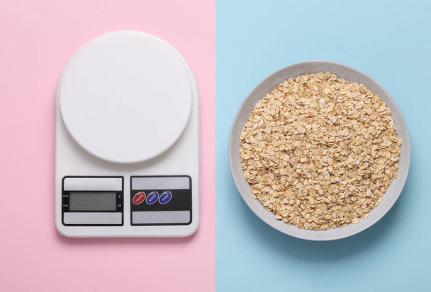 Calorie counting, weight loss, meal preparation, healthy eating concept. Electronic kitchen scales with a plate of raw oatmeal on pink blue background - Photo, Image
