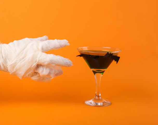 La mano della mamma avvolta in bendaggi bianchi raggiunge per un bicchiere di cocktail su uno sfondo arancione. Halloween partito concetto. - Foto, immagini