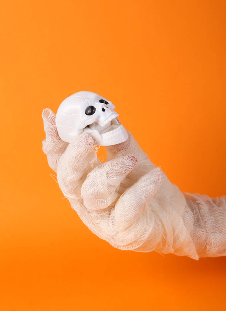 Maminčina ruka zabalená v bílých obvazech drží lebku na oranžovém pozadí. Halloween koncept - Fotografie, Obrázek