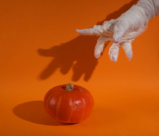 Mumiens Hand in weiße Binden gewickelt mit Kürbis auf orangefarbenem Hintergrund mit Schatten. Frohes Halloween. Süßes oder Saures - Foto, Bild