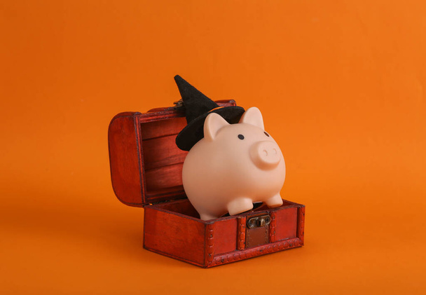 Halloween still life. Piggy banks with a witch's hat in a chest, orange background - Photo, Image