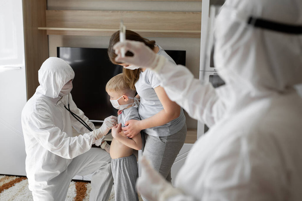Pediatrician doctor examining sick child in face mask and a protective suit. Sick boy with his mother at home. Kids home treatment of virus. Coronavirus pandemic. Covid-19 outbreak. - Fotografie, Obrázek