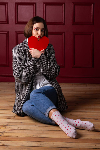 Short-haired woman in jacket sitting on wooden floor and red background holding red heart shape in front of her chin - Photo, Image