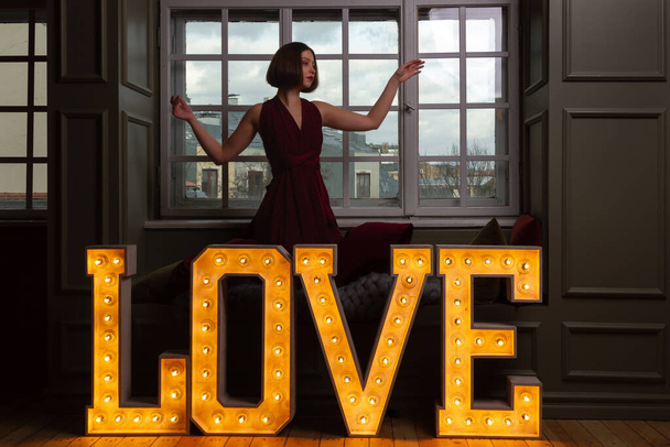 Una mujer con las manos en el aire en un vestido rojo detrás de una palabra AMOR hecho de letras brillantes - Foto, Imagen