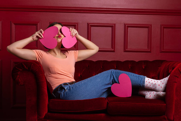 Women in jeans with lips shaping kiss on a couch holding heart-shaped paper sheets in front of her face - Photo, Image