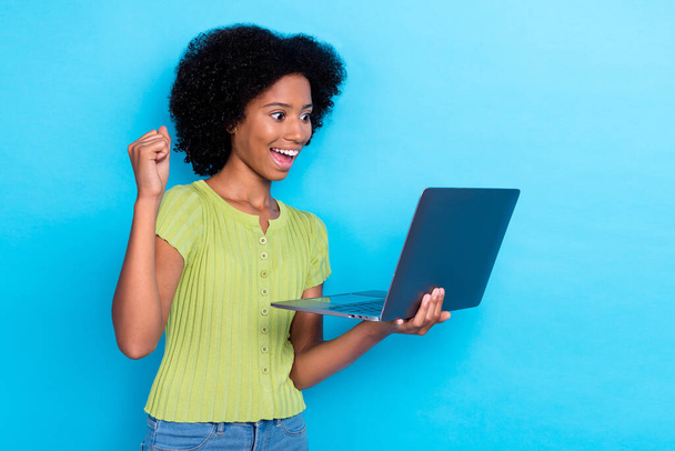 Portrait of ecstatic overjoyed girl with wavy hairdo dressed green t-shirt look at laptop clench fist up isolated on blue color background. - Fotoğraf, Görsel