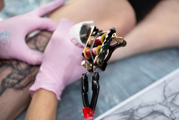 Close-up of the tattoo machine. A tattoo. A male master in a mask and gloves creates a pattern on the skin of a woman. - Photo, image