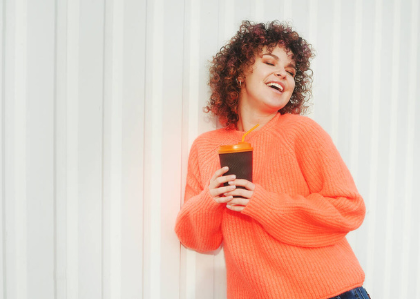 Smiling young woman with curly hair holds cup with coffee outdoors - Fotó, kép