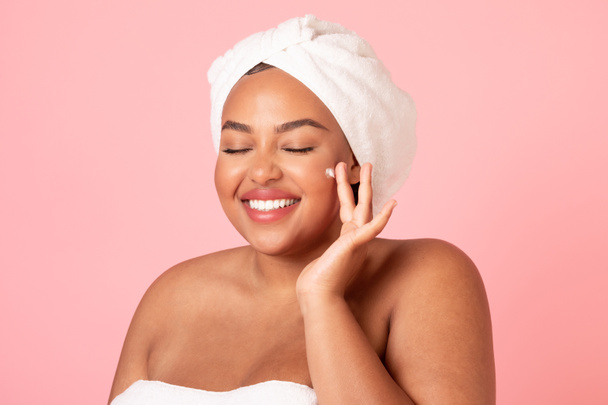 Happy black body positive woman applying beauty product on her face and smiling, lady wrapped in towel using face cream after shower, isolated on pink studio background - Foto, imagen