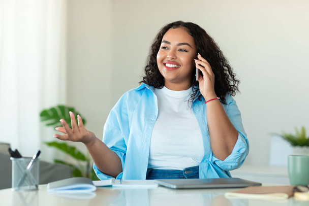 Retrato de la mujer afroamericana feliz de gran tamaño hablando en el teléfono inteligente, sentado en el escritorio en casa, espacio libre. Emocionado negro cuerpo positivo dama teniendo conversación telefónica - Foto, imagen