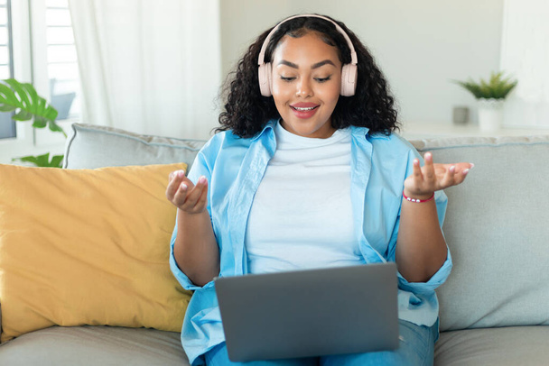 Видеоконференция. Портрет положительной черной пухлой женщины в беспроводной гарнитуре с веб-звонком через ноутбук, сидящей на диване, говорящей и жестикулирующей на веб-камеру, копирующей пространство - Фото, изображение
